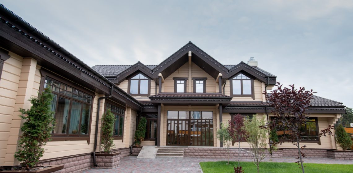 A two-story beige house with dark trim and gabled roof, surrounded by landscaped greenery and a paved walkway.