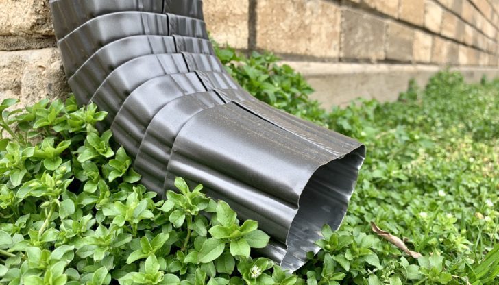 A damaged black plastic gutter lies partially buried in green foliage against a stone wall.