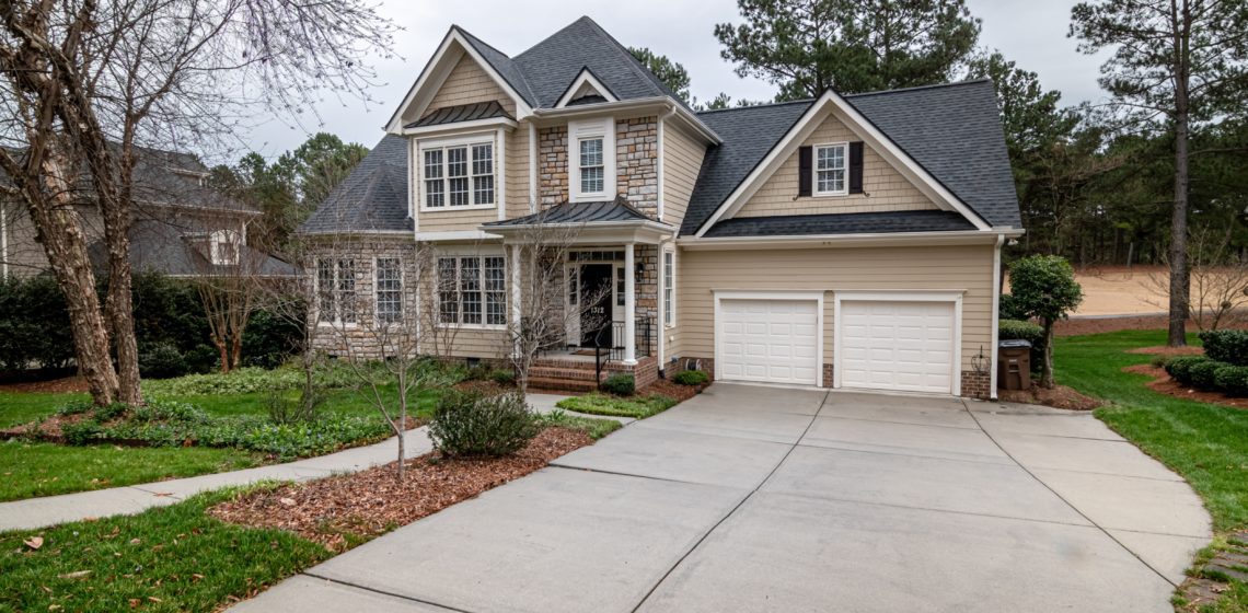 A beige two-story house with stone accents and a double garage, surrounded by a landscaped yard and mature trees.