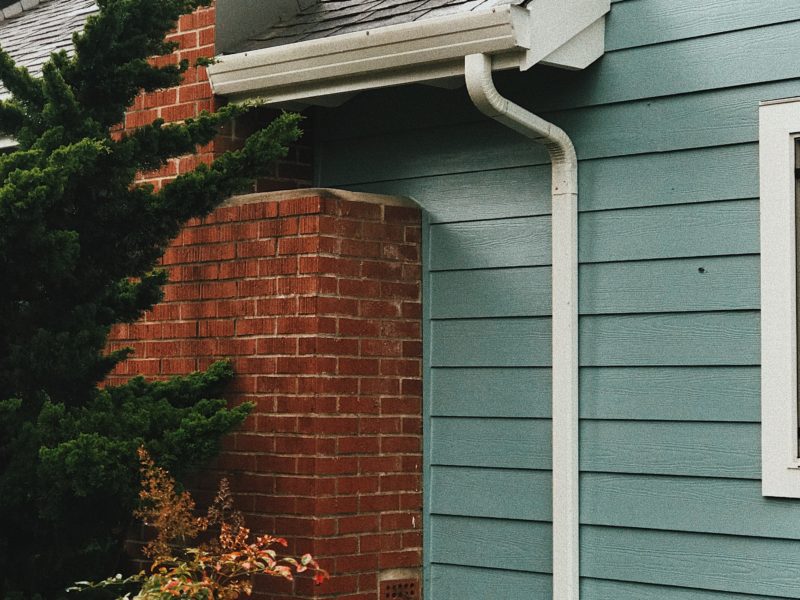 A close-up of a house corner with red brick and teal siding, featuring a white gutter and downspout.
